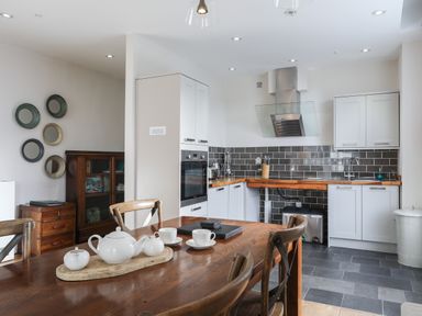 A kitchen with white cabinets a wooden table with a white tea set and wooden chairs at Harlech Apartments - Bendigeidfran in Harlech