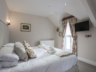 A bedroom with a bed with pillows and rolled towels a window with patterned curtains a wall mounted tv and a framed picture at Harlech Apartments - Bendigeidfran in Harlech