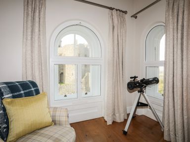 A corner room with a plaid armchair a telescope on a tripod and arched windows showing a castle outside at Harlech Apartments - Gwern in Harlech