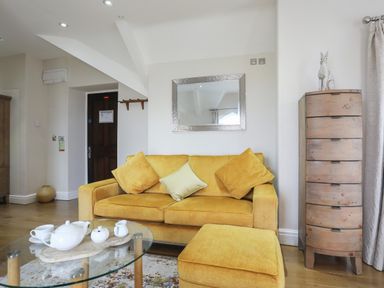 A living room with a yellow sofa and ottoman a glass coffee table with tea set a tall wooden chest and a mirror on the wall at Harlech Apartments - Gwern in Harlech