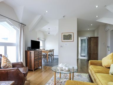 A living room with a glass coffee table a yellow sofa a brown leather armchair a wooden chest of drawers and a dining area in the background at Harlech Apartments - Gwern in Harlech