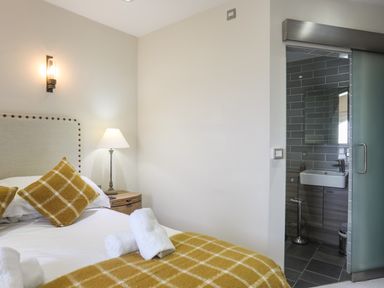 A bedroom with a bed with yellow checkered pillows and blanket and a bedside table with a lamp next to a bathroom with gray tiles and a sliding door at Harlech Apartments - Gwern in Harlech