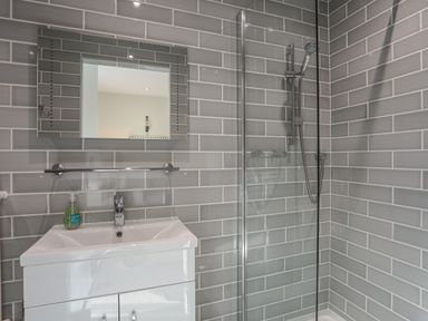 A bathroom with a sink cabinet, wall-mounted mirror, hand soap, glass shower enclosure, and a showerhead at Harlech Apartments - Matholwch in Harlech