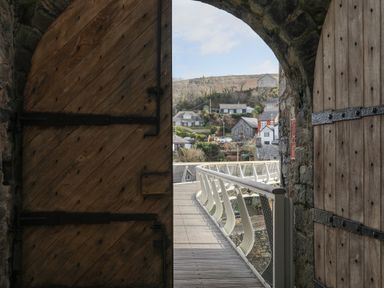 A wooden door opening to a curved walkway with railings overlooking a village with houses and hills at Harlech Apartments - Matholwch in Harlech