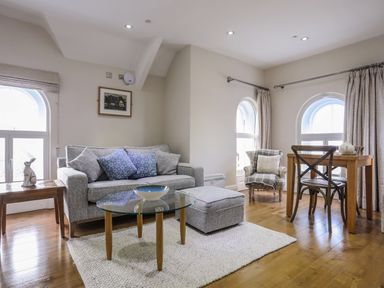 A living area with a grey sofa, patterned cushions, a glass coffee table, an armchair, a wooden dining table and chairs at Harlech Apartments - Matholwch in Harlech