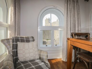 A room with a plaid armchair covered with a blanket and pillow next to a wooden table and chair by an arched window showing a castle outside at Harlech Apartments - Matholwch in Harlech