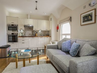 A kitchen with white cabinets and appliances next to a living area with a gray sofa and glass coffee table at Harlech Apartments - Matholwch in Harlech