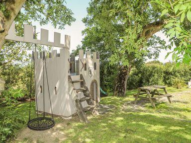 An outdoor play area with a wooden castle structure a tire swing hanging from a tree and a wooden picnic table on grass at Hen Gilfach in Llanfairyngnhornwy