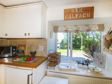 A kitchen corner with white cabinets a microwave onions and peppers on the wooden counter a window with a floral blind and a view of a garden at Hen Gilfach in Llanfairyngnhornwy