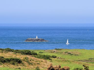 A coastal landscape with grassy fields in the foreground a small rocky island with a white structure and a sailboat on the sea at Hen Gilfach in Llanfairyngnhornwy