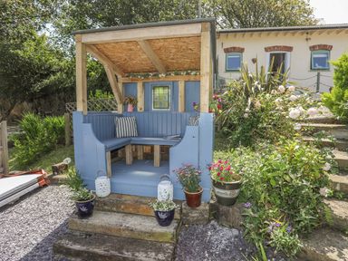 An outdoor wooden seating area painted blue with cushions and flower pots surrounded by plants and stone steps at Hen Gilfach Stable in Llanfairyngnhornwy