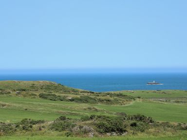 A coastal landscape with green grassy fields and shrubs overlooking the blue ocean with a small island and a lighthouse at Hen Gilfach Stable in Llanfairyngnhornwy