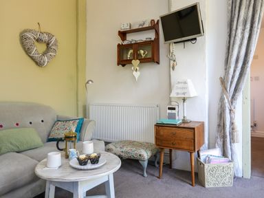 A living room with a gray sofa and cushions a round wooden coffee table with a teapot and cupcakes a wooden side table with a lamp and radio a small floral footstool a wall-mounted TV and shelf with decorations at Hen Gilfach Stable in Llanfairyngnhornwy