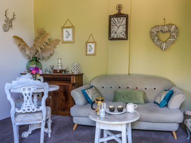 A living room with a gray sofa, round white table with muffins and mugs, decorative wall clock, heart-shaped wall decor, and a white chair at Hen Gilfach Stable in Llanfairyngnhornwy