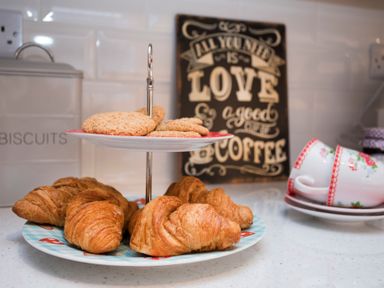 A kitchen counter with croissants and cookies on a tiered plate and stacked teacups and saucers by a biscuit tin at Hirfron in Llanberis