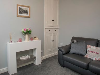 A small living room corner with a black leather sofa with cushions and a white decorative fireplace with flowers at Hirfron in Llanberis