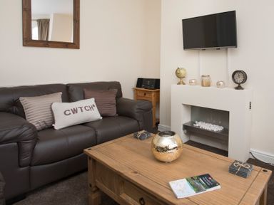 A living room with a brown leather sofa with cushions a wooden coffee table with a decorative item and book a wall-mounted TV and a decorative fireplace at Hirfron in Llanberis