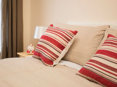 A bed with beige pillows and red striped cushions next to a bedside table with a lamp and a small decorative item at Hirfron in Llanberis