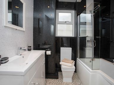 A bathroom with a white sink vanity a toilet with towels on top and a bathtub with a glass shower screen at Hirfron in Llanberis