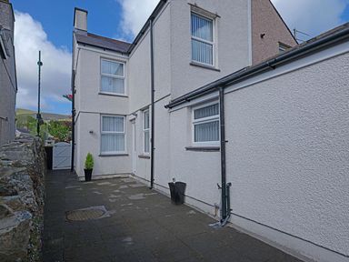 An outdoor paved area beside a white textured house with windows a potted plant and a stone wall at Hirfron in Llanberis