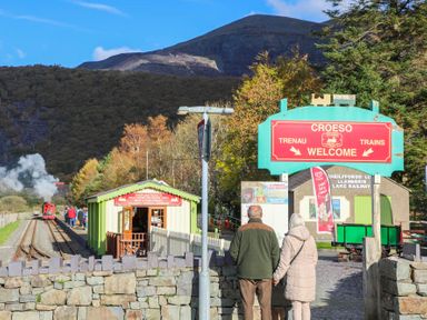 Two people standing in front of a railway station platform with a sign welcoming trains at Hirfron in Llanberis