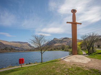 A large sword sculpture on a grassy bank beside a lake with mountains and trees in the background at Hirfron in Llanberis