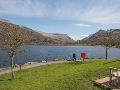 A lakeside path with two people walking near mountains and trees at Hirfron in Llanberis