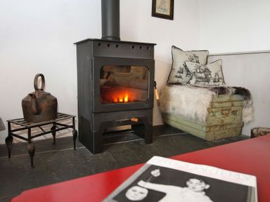 A room with a woodburning stove a metal kettle on a small table and a cushioned bench with fur throw and ship patterned pillows at Big House in Llandecwyn