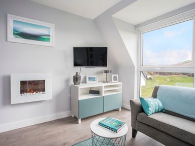 A living room with a wall-mounted TV a fireplace a gray sofa with blue cushions a sideboard and a round coffee table at Isallt in Trearddur Bay