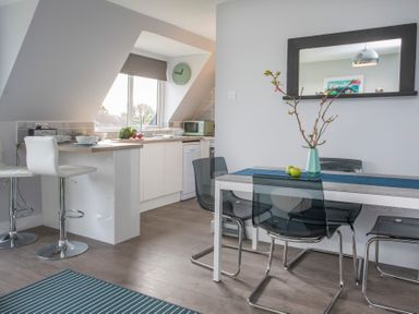 A kitchen and dining area with white cabinets, bar stools, a dining table, chairs, a mirror, and a vase with branches at Isallt Trearddur Bay