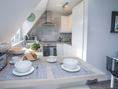 A kitchen with white cabinets gray tiled backsplash a stove and a breakfast bar set with bowls cups and bread at Isallt in Trearddur Bay