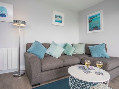 A corner sofa with blue and green cushions a floor lamp and a round coffee table with two glasses and magazines at Isallt in Trearddur Bay