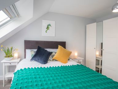 A bedroom with a bed with cushions and a green blanket two bedside tables with lamps a wardrobe with mirror and a skylight window at Isallt in Trearddur Bay