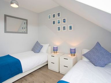 A bedroom with two single beds blue pillows and blankets two white nightstands with blue lamps and framed pictures on the wall at Isallt in Trearddur Bay