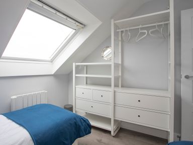 A bedroom with a bed covered by a blue blanket a skylight window and a white wardrobe with drawers and hangers at Isallt Trearddur Bay