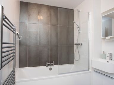 A bathroom with a white bathtub shower a glass shower screen a gray tiled wall a white sink with a mirror and a chrome towel rail at Isallt in Trearddur Bay