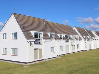 A row of white townhouses with brown roofs and several windows facing a grassy lawn at Isallt Trearddur Bay