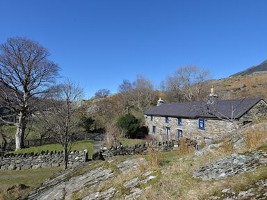 A stone house with blue window frames surrounded by trees and stone walls under a clear blue sky at Llwyn Bedw in Nant Peris