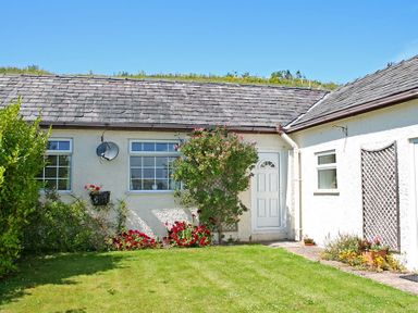 The exterior of a cottage with a grass lawn flowers around the walls and a white door at Mermaid Cottage in Brynsiencyn