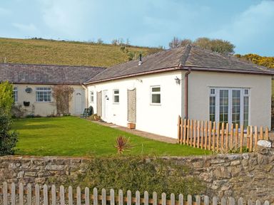 A single story house with white walls and a garden with grass and a wooden fence at Mermaid Cottage in Brynsiencyn