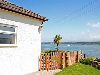Exterior of a white building with a wooden fence and gate overlooking a body of water with boats and a palm tree at Mermaid Cottage in Brynsiencyn
