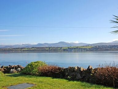 A view of a calm body of water with greenery and rocks in the foreground and hills and houses in the background at Mermaid Cottage in Brynsiencyn
