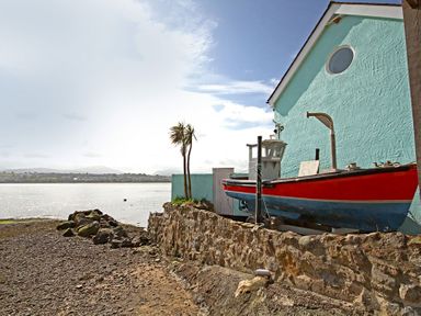 A red and blue boat next to a turquoise building by a rocky shore with water and distant hills at Mermaid Cottage in Brynsiencyn