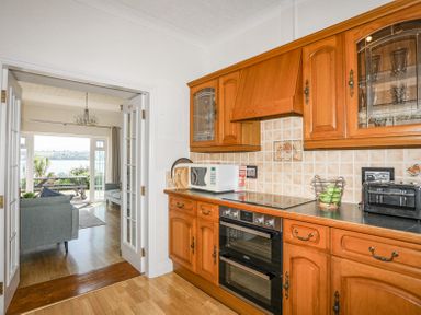 A kitchen with wooden cabinets a double oven microwave and toaster leading to a living room with sofas and a large window with a view at Mermaid Cottage in Brynsiencyn