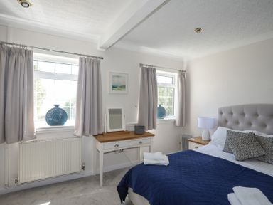 A bedroom with two windows grey curtains a bed with blue quilt and pillows a bedside table with lamp and a dressing table with mirror at Mermaid Cottage in Brynsiencyn