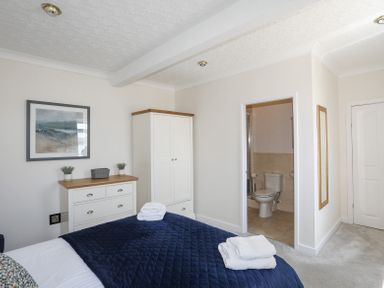 A bedroom with a bed covered in a dark blue quilt facing a white wardrobe and chest of drawers with plants and artwork on the wall at Mermaid Cottage in Brynsiencyn