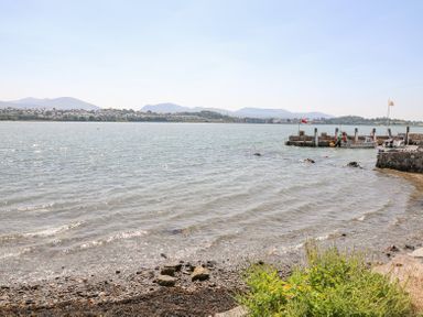 A view of a rocky shoreline with water small boats docked and hills in the background at Mermaid Cottage in Brynsiencyn