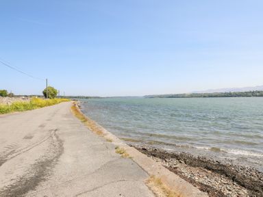 A coastal road alongside a body of water with vegetation and trees in the distance at Mermaid Cottage in Brynsiencyn