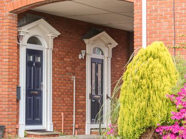 Two black entrance doors on a red brick building with white trim under a covered entryway with green shrub and pink flowers
