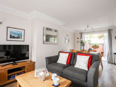 A living room with a gray sofa and wooden table facing a TV and a dining area with open doors to a garden at Mews Cottage in Menai Bridge